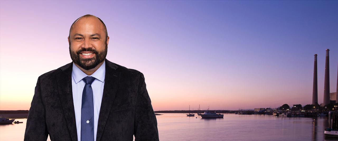 Person standing outdoors in front of a harbor at sunset.