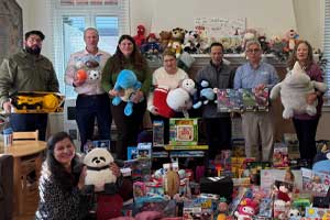 Group of people standing indoors behind a table filled with wrapped gifts, toys, and stuffed animals.