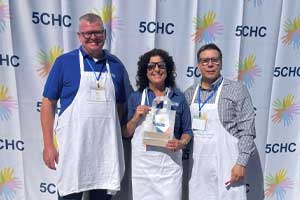 Three people standing outdoors wearing matching aprons, holding items in front of a branded event backdrop.