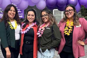 Group of people standing outdoors together wearing event shirts and leis beneath purple balloons.