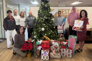 Group of people standing indoors around a decorated holiday tree and an arrangement of wrapped gifts and poinsettias.