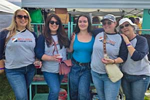 Group of people standing together outdoors near a table at a community event, with tents and equipment visible in the background.