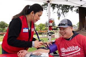 Two people seated outdoors at an event table, with one person using a blood pressure cuff on the other, under a canopy tent.