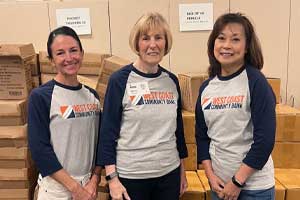 Three people standing indoors in front of stacked cardboard boxes, wearing matching shirts at a volunteer workspace.