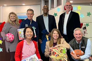 Group of people standing and seated indoors holding wrapped gifts and items in front of a wall decorated with colorful star cutouts.