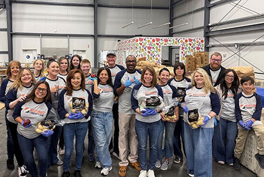 Group of volunteers in a warehouse holding bagged food items, wearing matching shirts and gloves at a food bank event