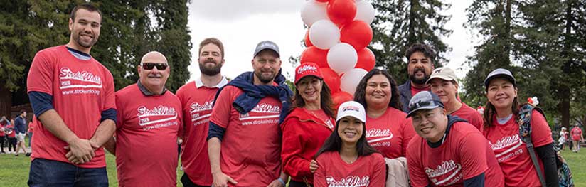Group of people standing together outdoors at a community walk event, wearing matching shirts and gathered near red and white balloons.