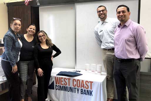 Group of people standing in a classroom in front of a whiteboard, with a West Coast
Community Bank table set up with cups, folders, and pens.