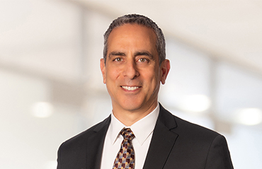 Professional headshot of a man in a suit and patterned tie against a softly blurred indoor background.
